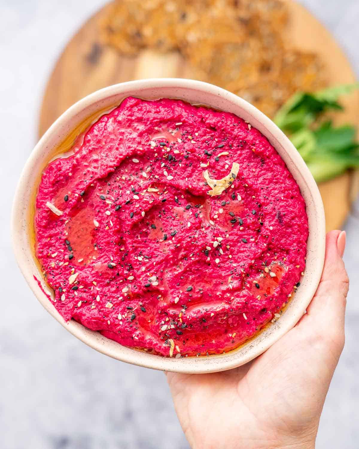 A hand holding up a bowl of beetroot hummus above a chopping board of green vegetables beneath it. 