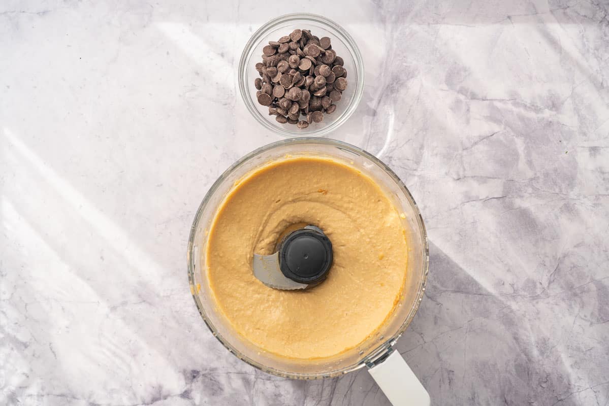 A food processor sitting on the bench with ingredients whizzed together inside of it, sitting next to a ramekin of chocolate chips 