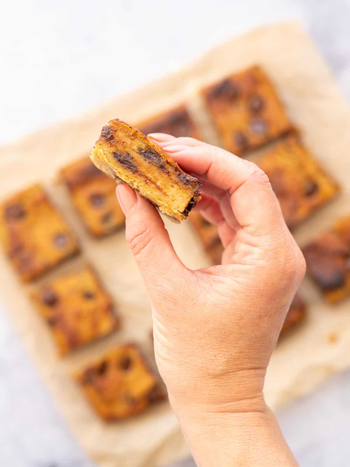 Baked Chickpea Blondies sliced and sitting on brown crinkled baking paper with a hand holding up one individual piece. 