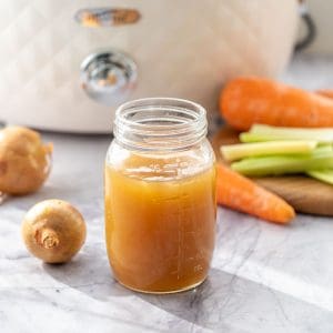 A glass jar of golden brown broth sitting in front of a slow cooker and vegetables on a marble bench top.
