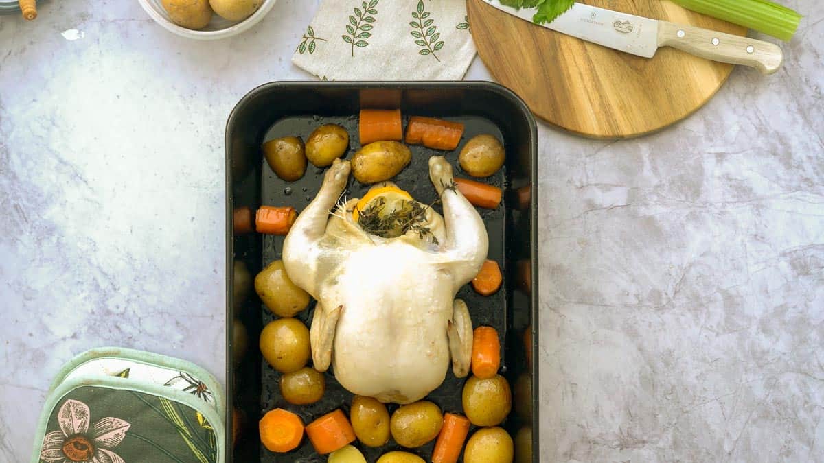 A large black roasting dish on a bench filled with roasted potatoes and carrots and a whole lemon and thyme stuffed chicken sitting amongst them.  Next to it, is a chopping board with a large knife and celery stick, a oven mit and a small bowl of potatoes. 