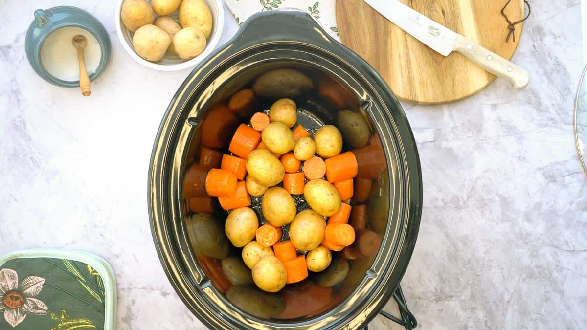 A large crockpot sitting on the bench, with baby potatoes and chopped carrots inside of it, sitting next to a jar of salt, a cutting board with a knife on it and a small bowl of potatoes. 