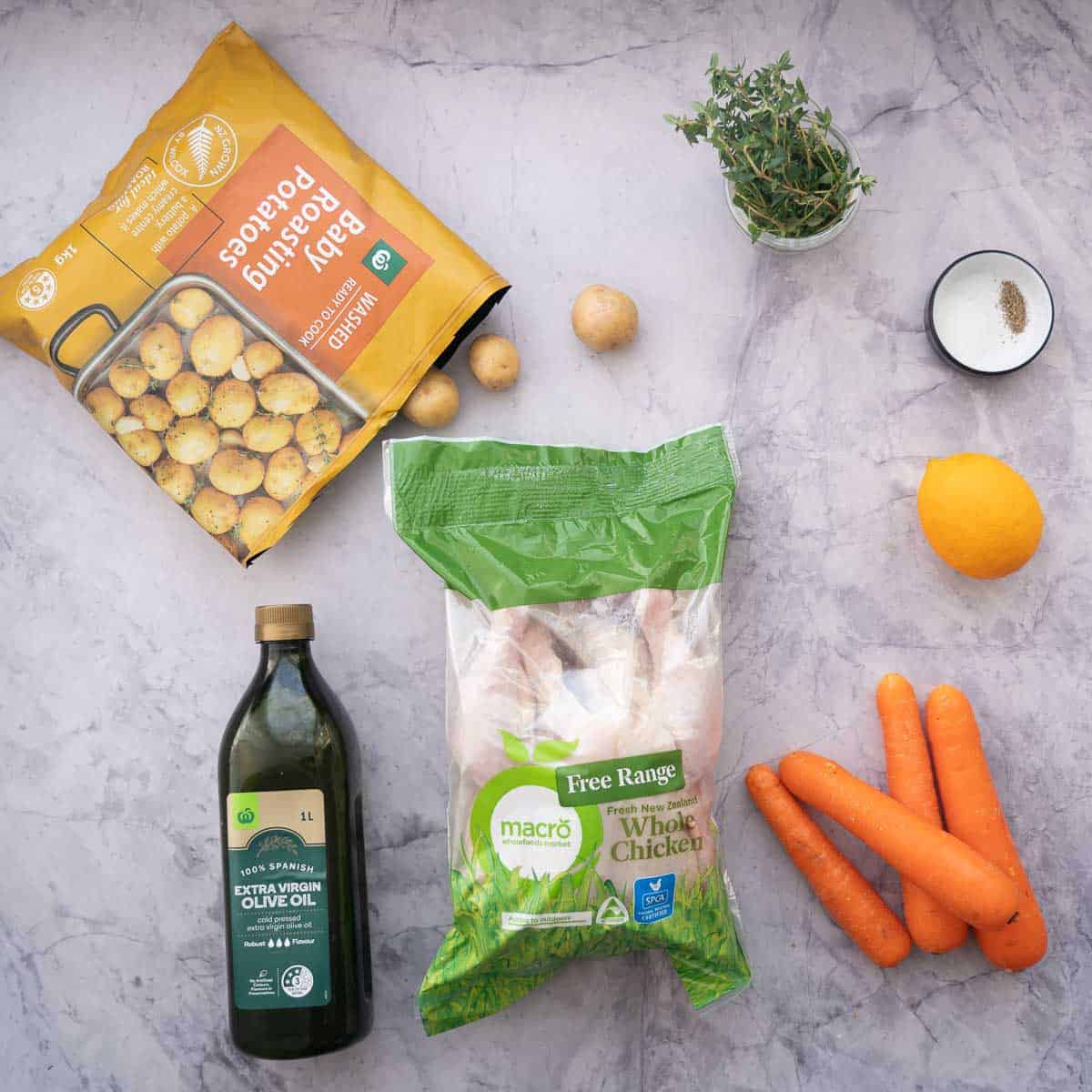 All the ingredients laid out on the bench to make Crocpot Roasted Chicken - A bottle of olive oil, a bag with a whole raw chicken inside of it. 4 carrots, a lemon and a bag of baby potatoes. 