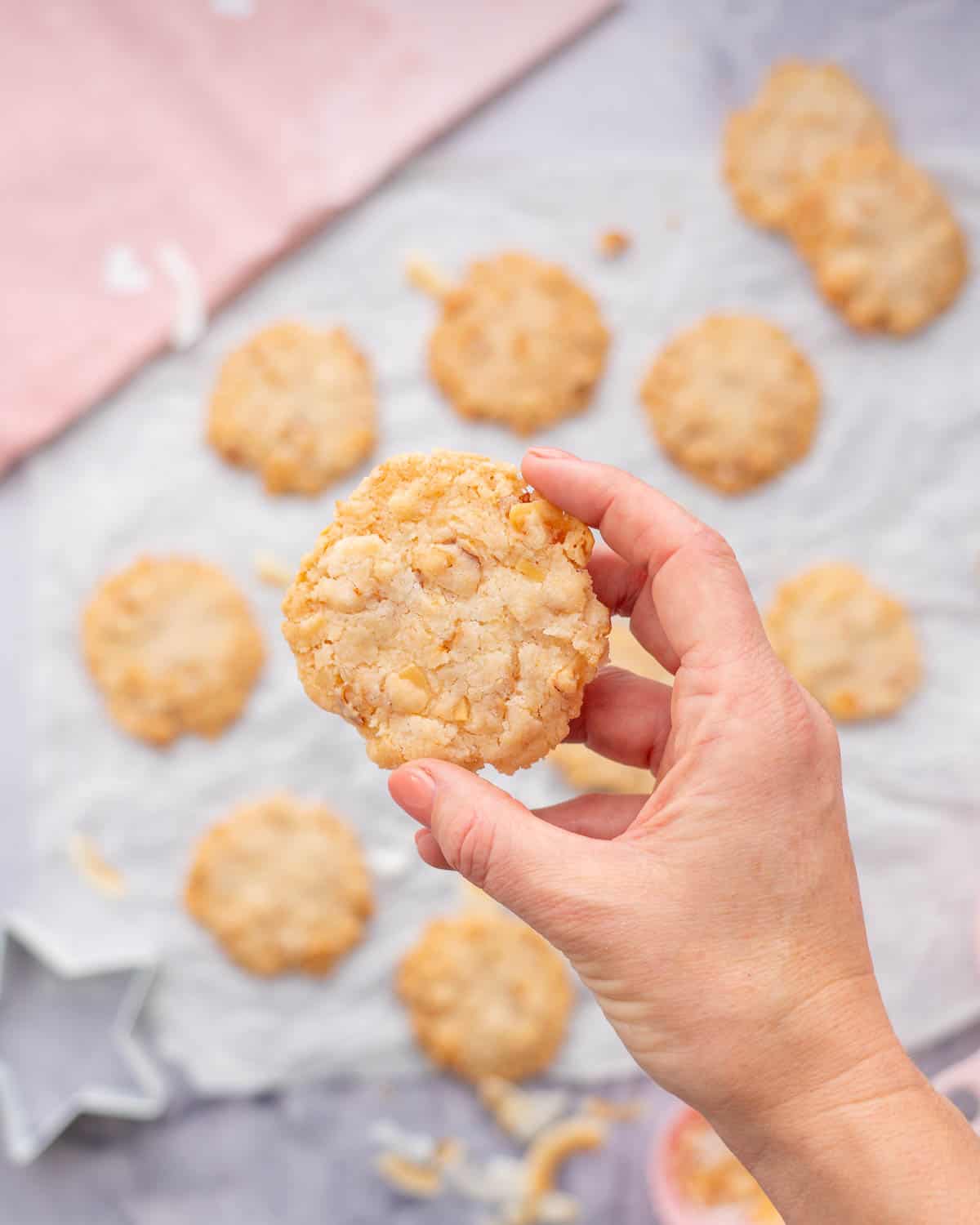 Crinkled baking paper with golden brown Gluten Free Coconut Cookies with a hand holding 1 cookie