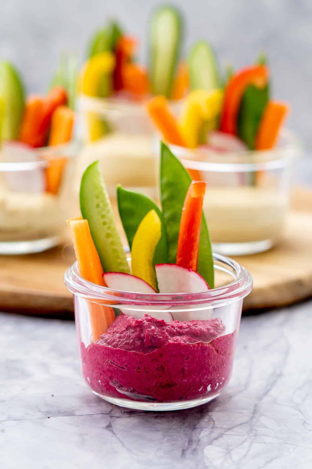Glass ramekins half filled with beetroot hummus and sliced pieces of radishes, carrots, sugar snap peas and cucumber sitting on the bench