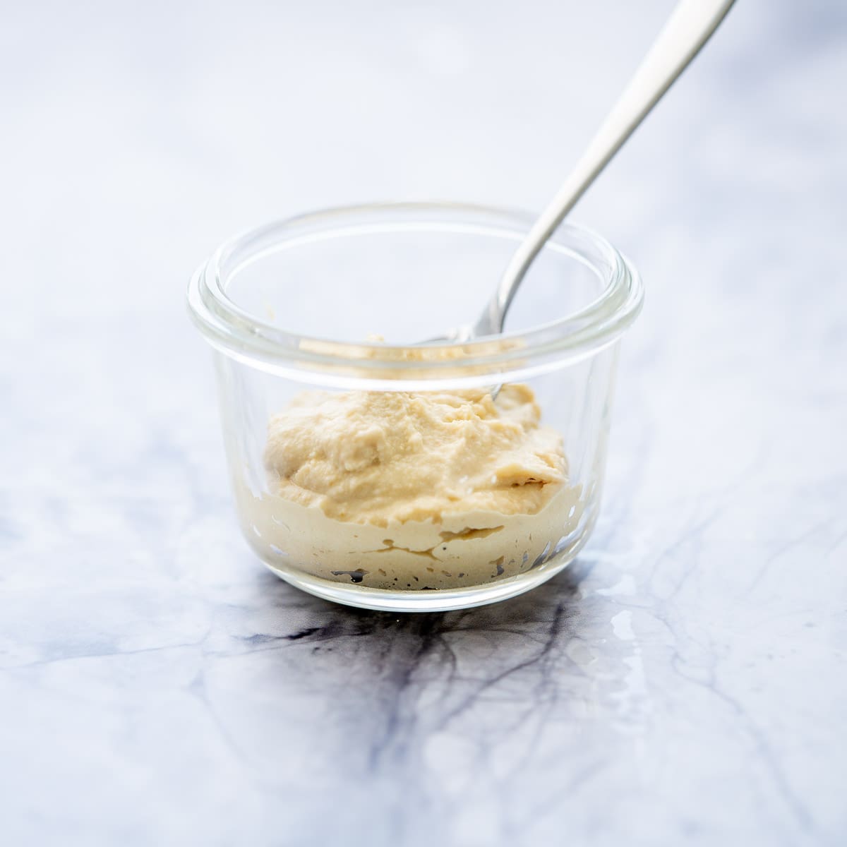 one glass ramekin half filled with hummus with a spoon  sitting on the bench with 