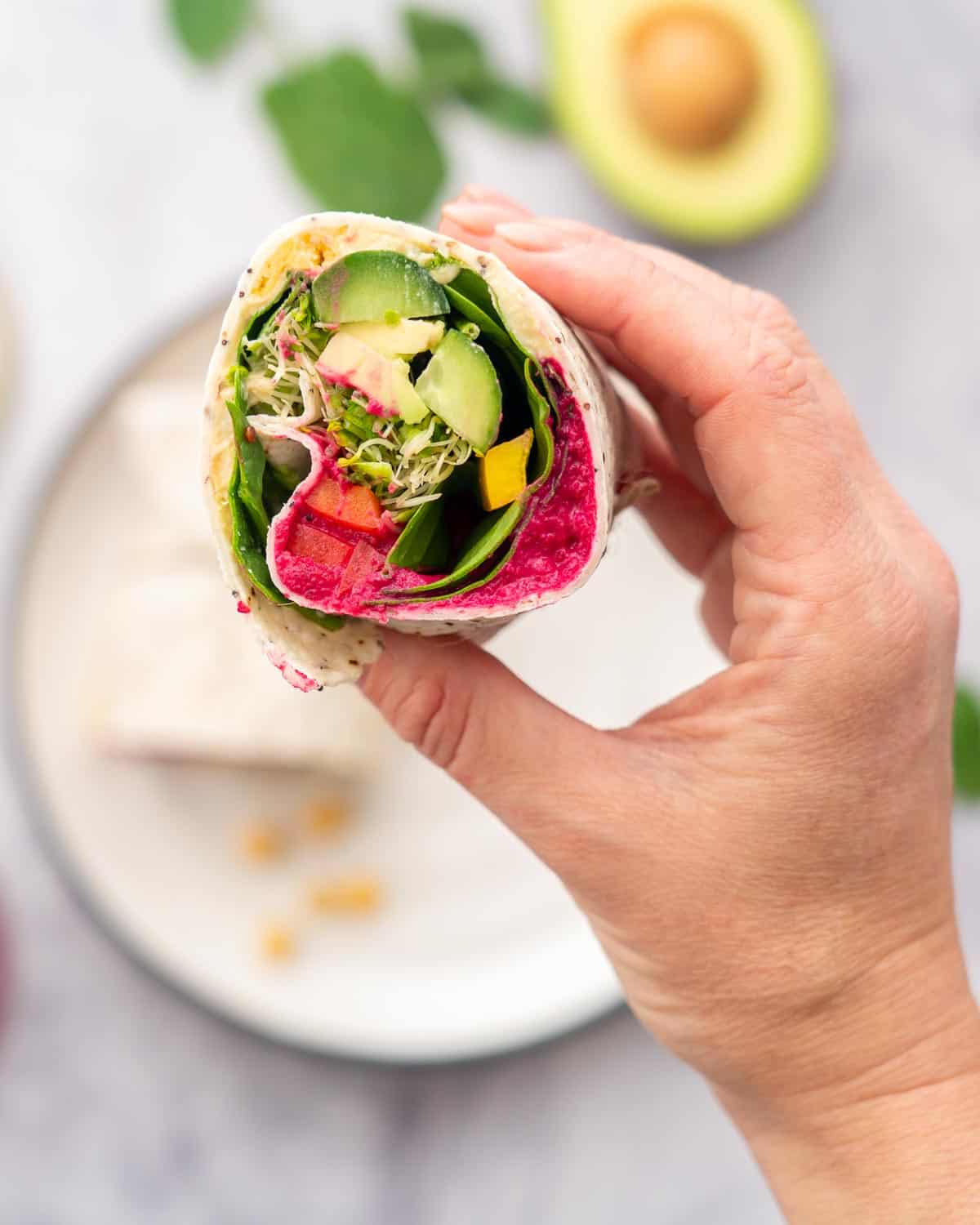 A hand holding up a halved filled wrap stuffed with beetroot hummus, cucumber, sprouts, spinach and capsicum