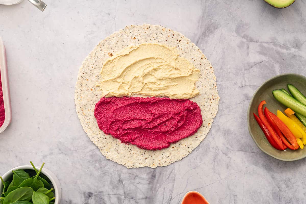A round wholegrain wrap spread with beetroot hummus and regular hummus, sitting next to individual ramekins of vegetables 