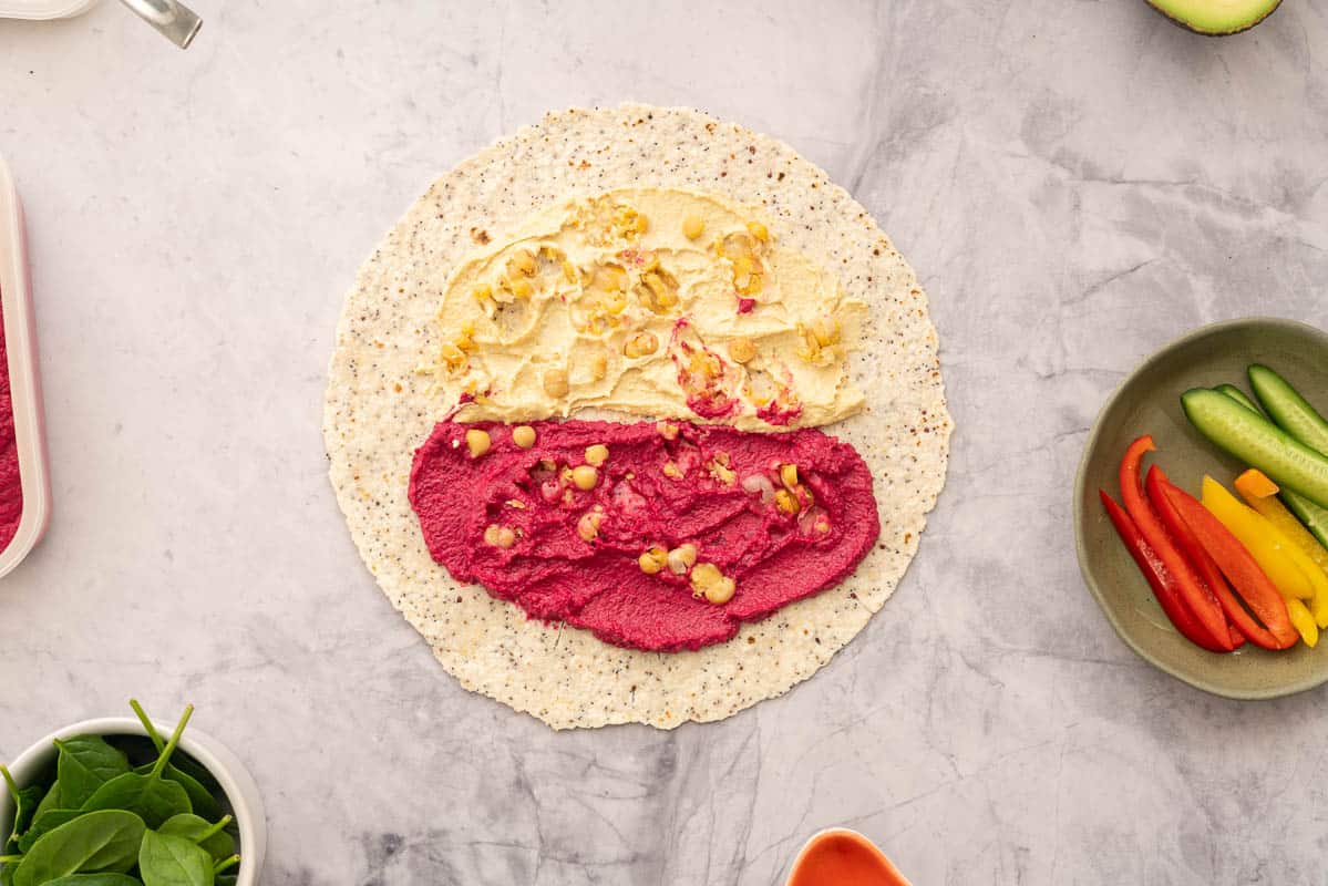A round wholegrain wrap spread with beetroot hummus and regular hummus, topped with chickpeas, sitting next to individual ramekins of vegetables 