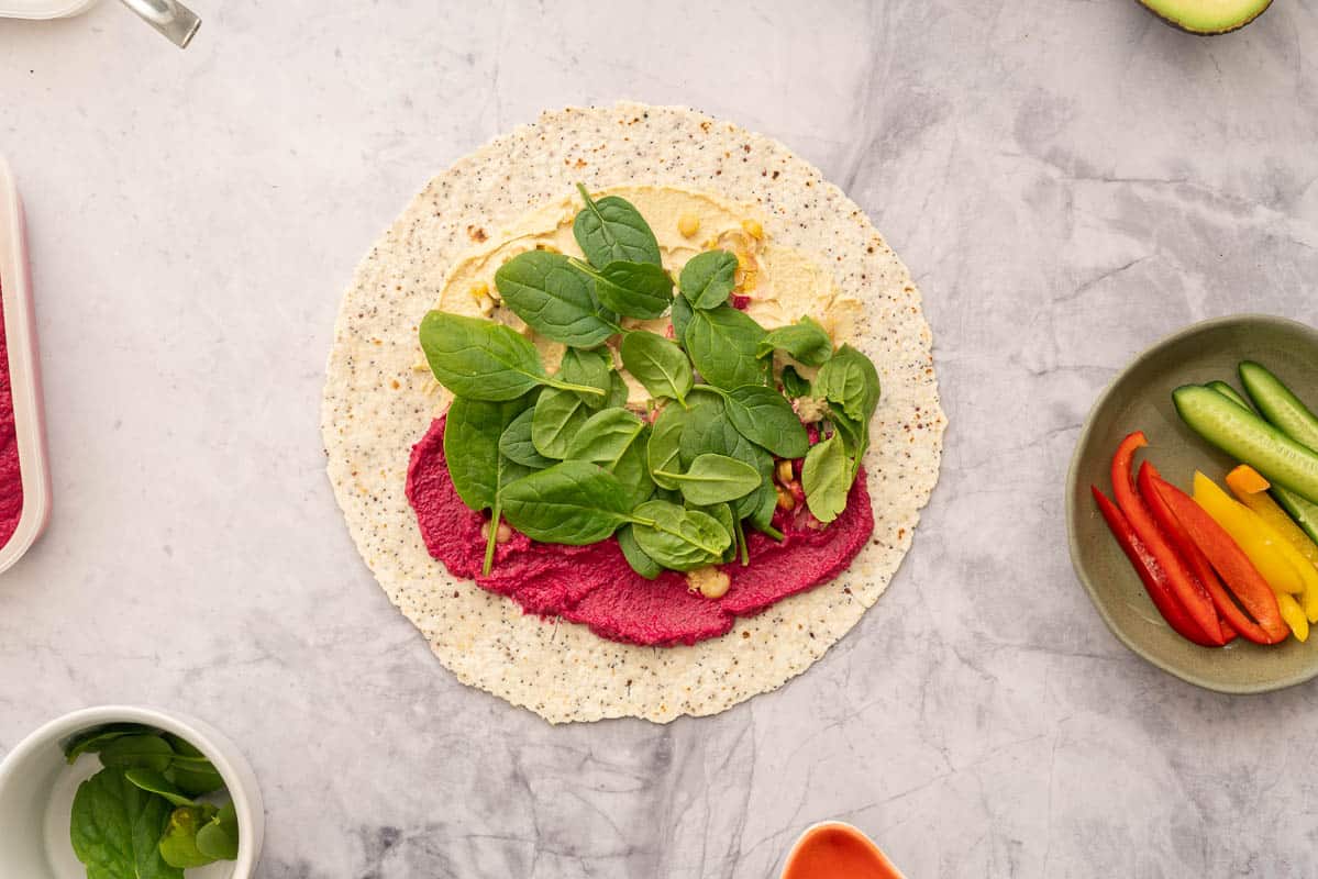 A round wholegrain wrap laid out on the bench  spread with beetroot hummus and regular hummus, topped with chickpeas and spinach sitting next to individual ramekins of vegetables 