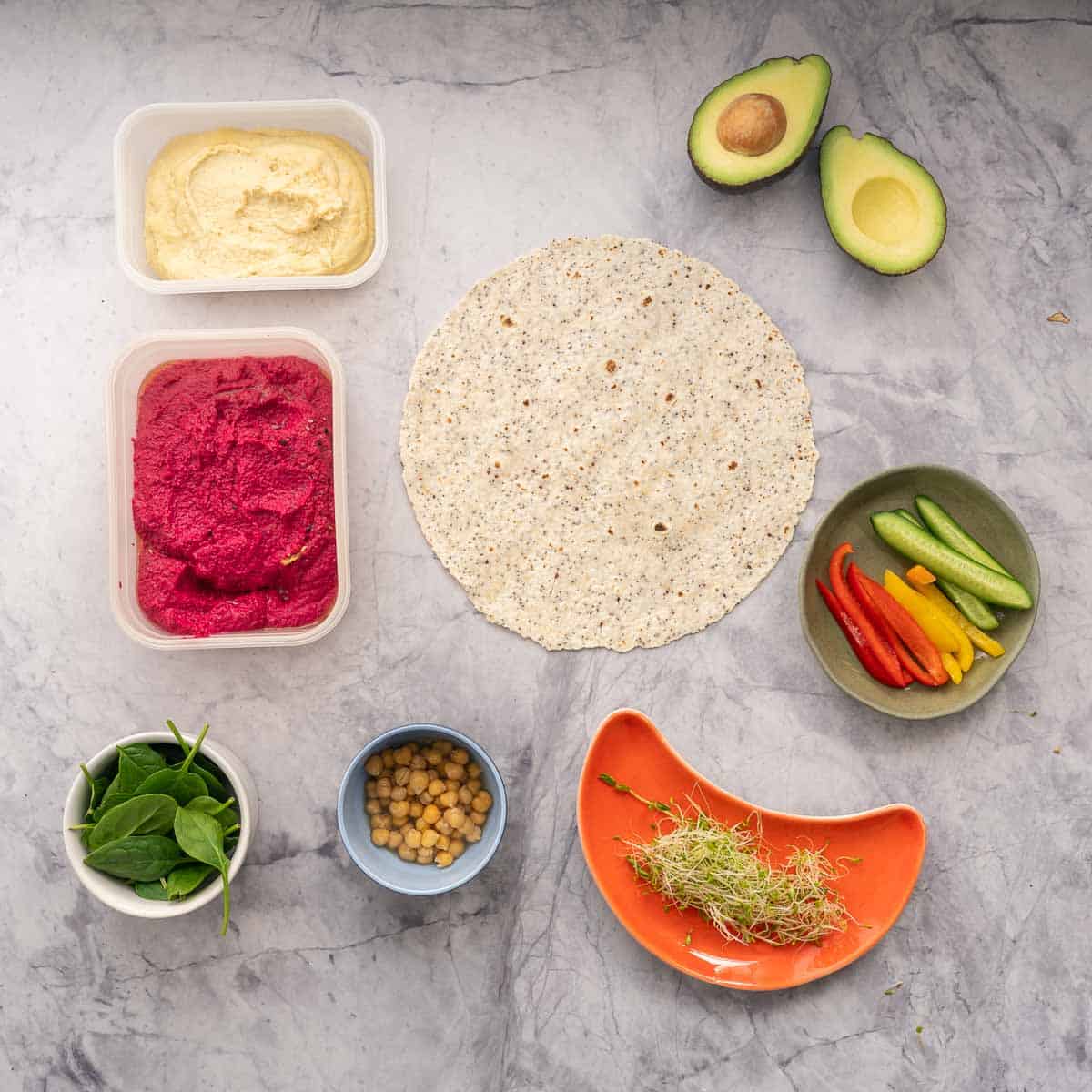 Containers of beetroot hummus and regular hummus sitting next to a wholegrain wrap, halved avocado, and individual ramekins of chickpeas, spinach, sprouts and sliced vegetables
