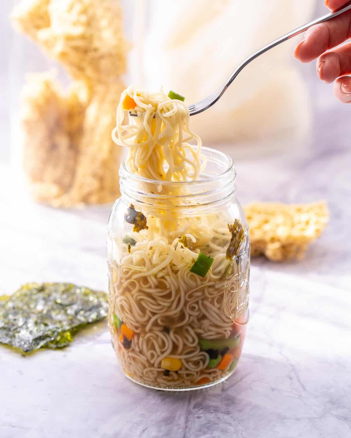 A mason jar on the bench with noodles, chopped carrot, corn kernels, beans and seaweed sheets teared up inside of it with a hand holding up a forkful or noodles