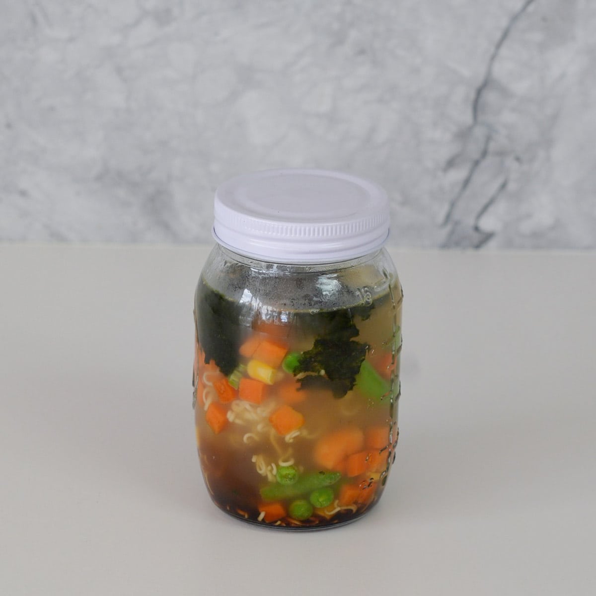 A mason jar with a lid on sitting on the bench with none broth, noodles, chopped carrot, corn kernels, beans and seaweed sheets teared up inside of it. 