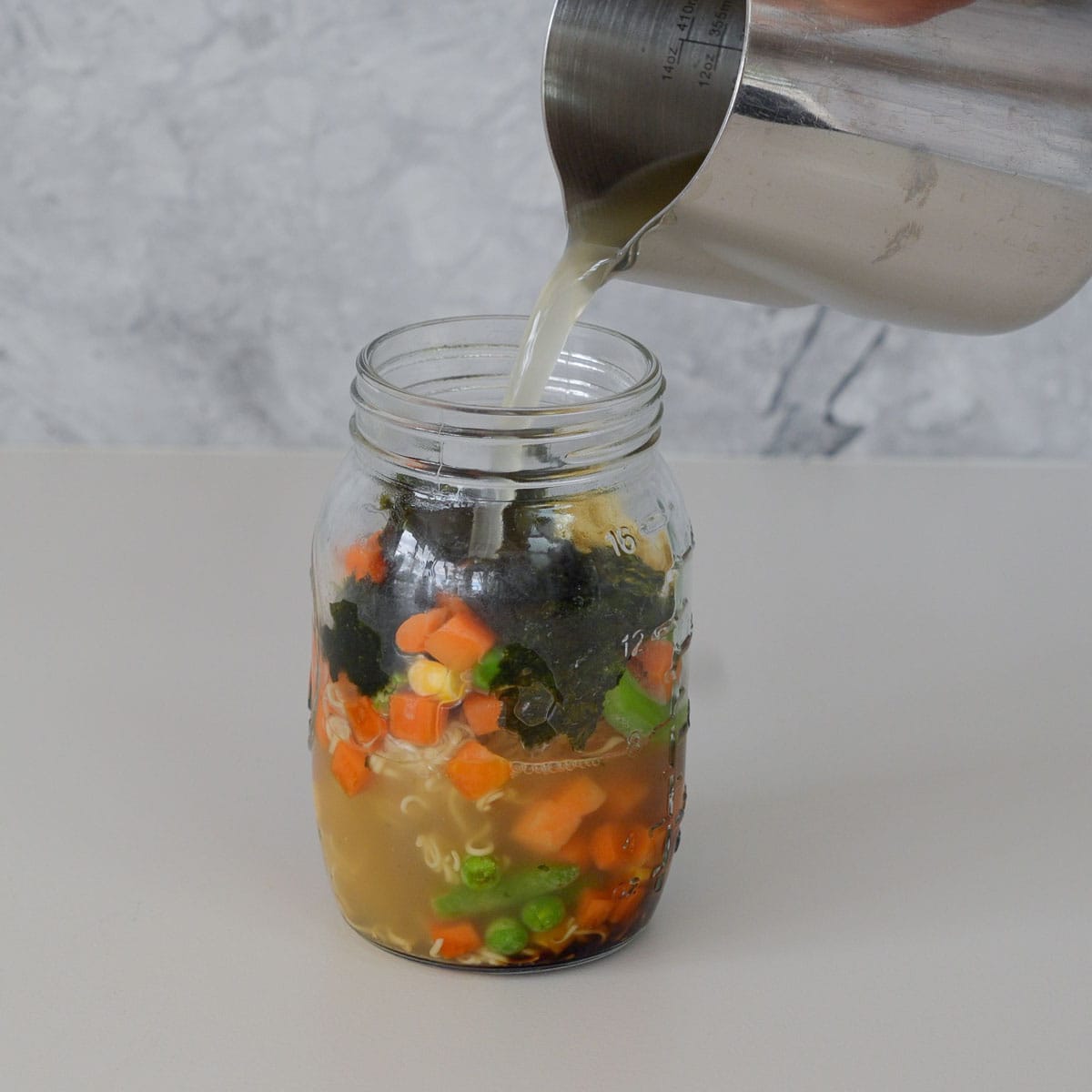 A mason jar on the bench with noodles, chopped carrot, corn kernels, beans and seaweed sheets teared up inside of it with a jug of bone broth being poured over the contents of the jar.