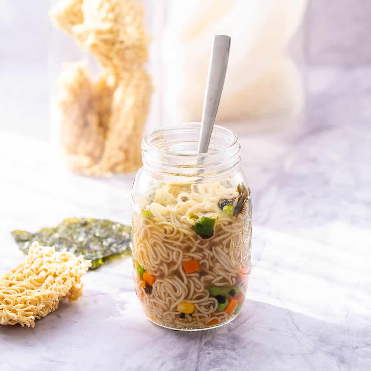 A mason jar on the bench with noodles, chopped carrot, corn kernels, beans and seaweed sheets teared up inside of it. 