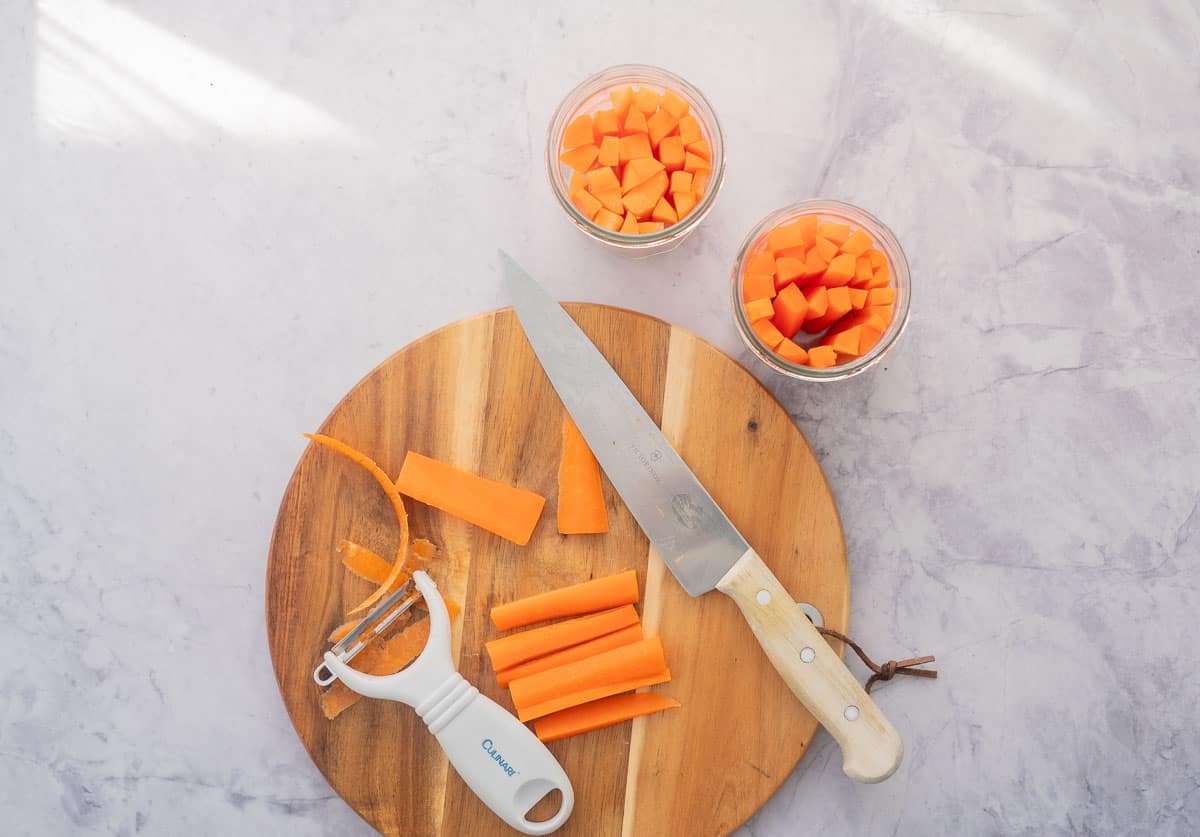 2 glass jars filled with battens of carrots, sitting next a chopping board  with sliced carrots a knife and a peeler on it. 