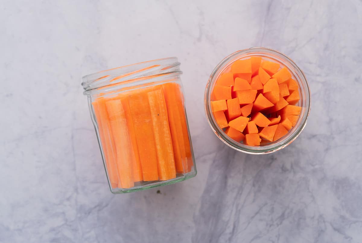 Two jars sitting on a bench with battened carrots inside of them, one turned on its side. 