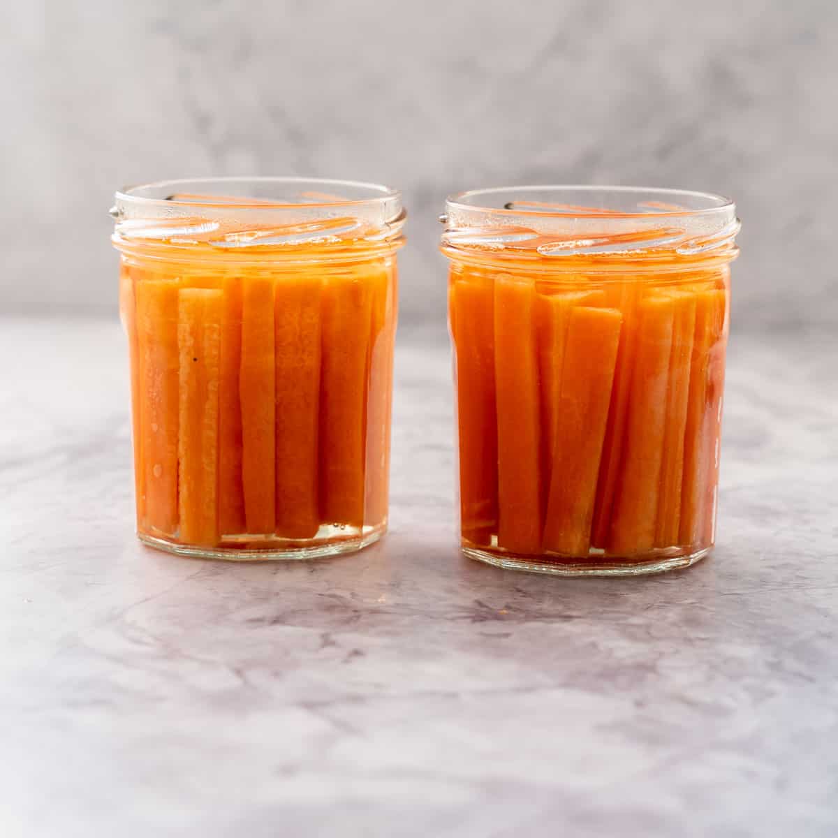 Two glass jars filled with battened carrots covered in pickling juice sitting on a bench 