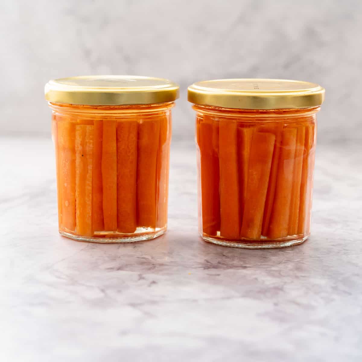 Two glass jars  with gold lids screwed on filled with battened carrots covered in pickling juice sitting on a bench