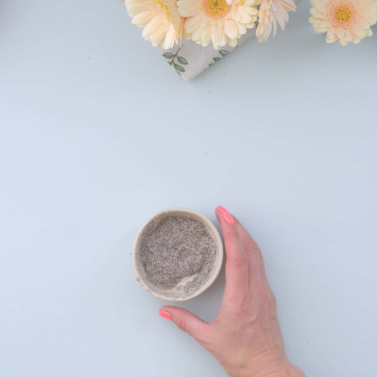 A hand reaching for a ramekin of soaked chia seed sitting on a light blue bench 