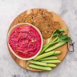 A bowl of pink hummus dip with vegetables anc crackers.