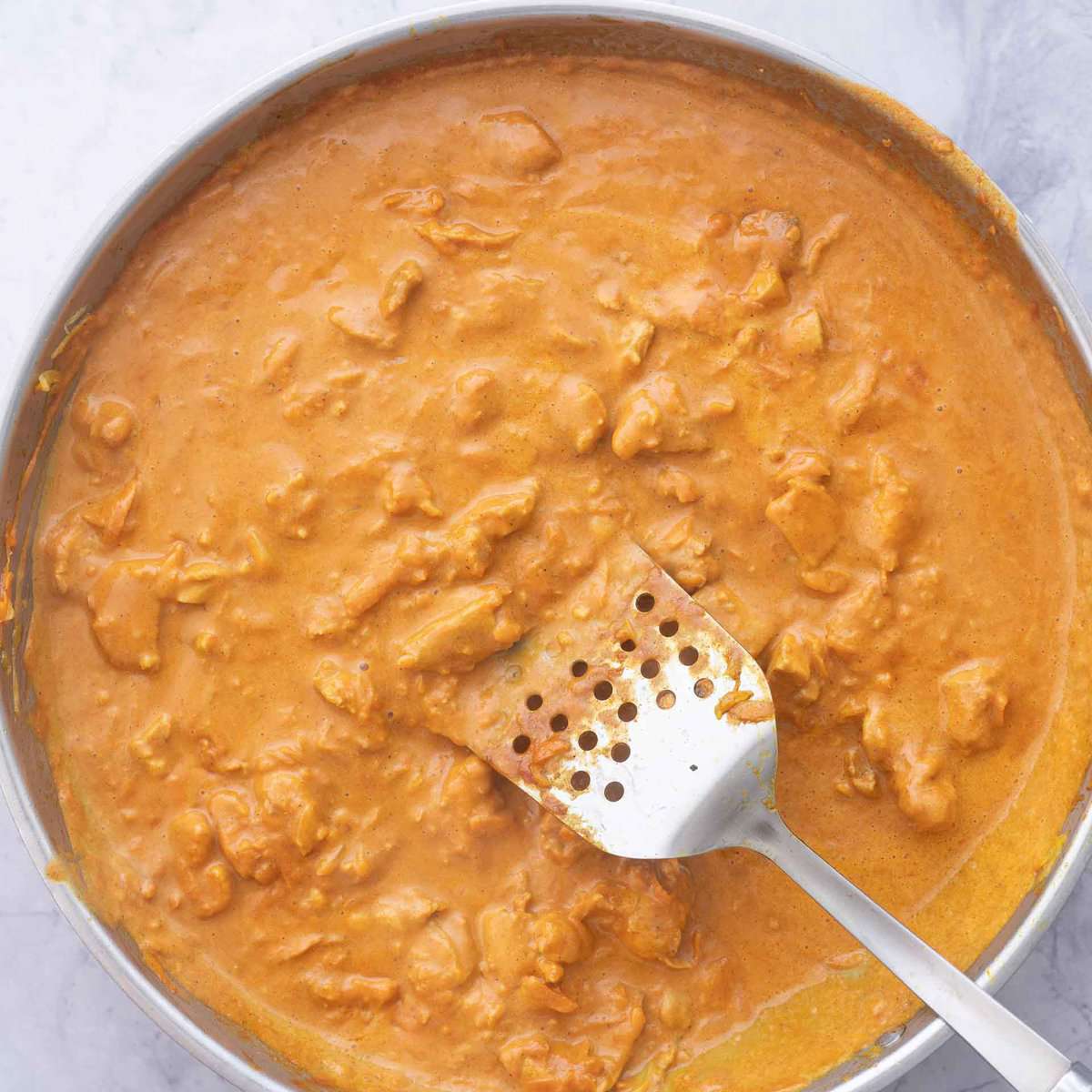 A pan full of cooked butter chicken with a  spatula resting on the side  