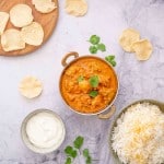 A copper curry bowl filled with butter chicken on a marble bench top with a bowl of rice, poppadoms and coriander leaves.