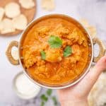 A bowl of butter chicken being held above poppadoms, rice and yogurt.