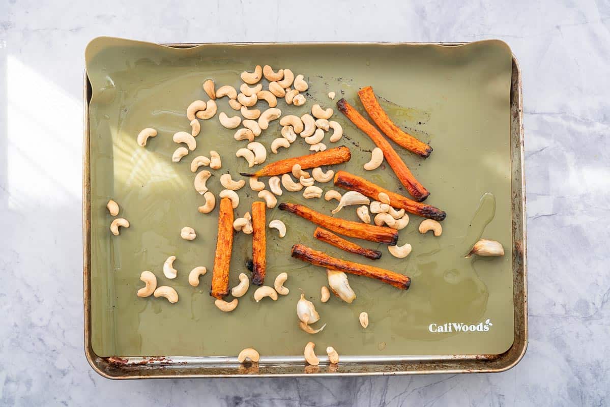A lined baking tray with roasted carrots, cashews and garlic cloves. 