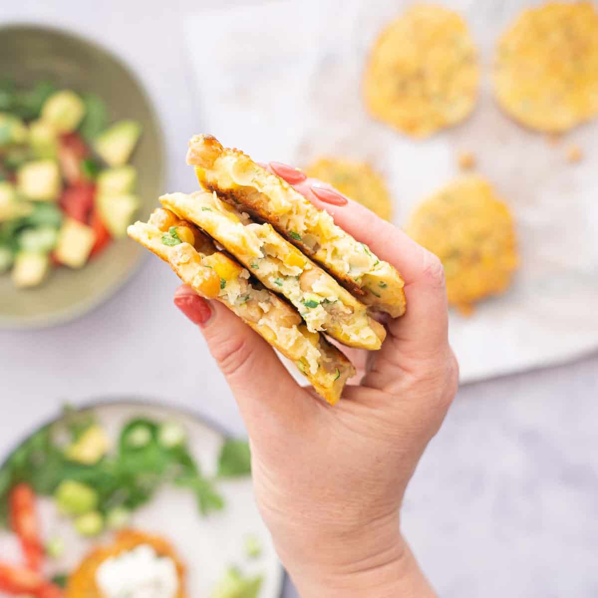 A hand holding three chickpea fritters above a bench of plates full of salads and more chickpea fritters