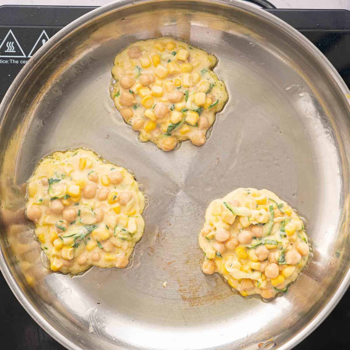 Three chickpea fritters cooking in an oiled frying pan 