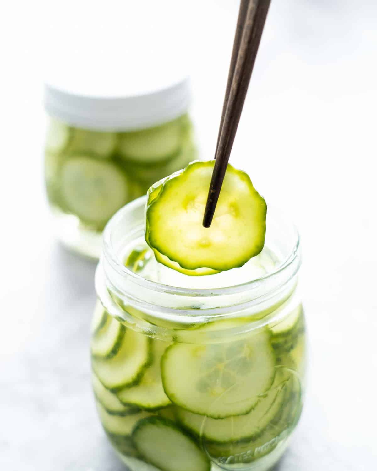 Two slices of cucumber being held with chopsticks above a jar of pickled cucumbers