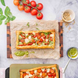 Two tomato tarts on a marble bench top surrounded by tomatoes, basil leaves and a small bowl of green pesto.