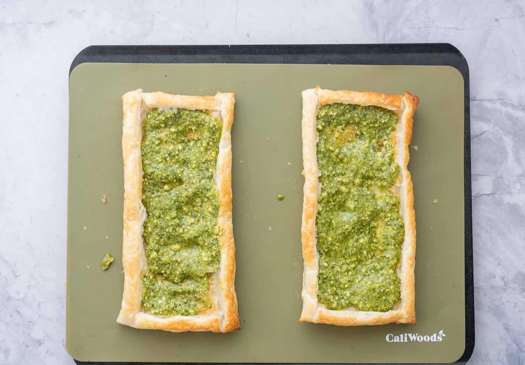 Two baked tarts on a baking tray with pesto spread across each of them. 