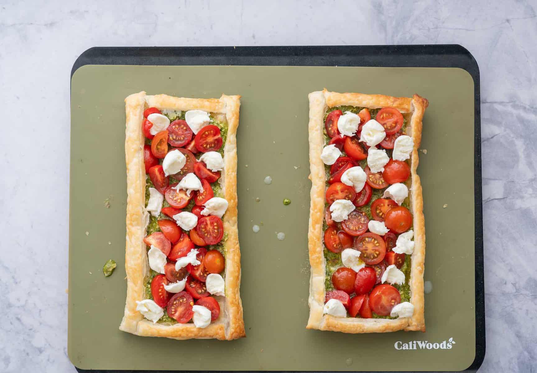 Two baked tarts on a baking tray with pesto spread across each of them a layered with sliced tomatoes and cheese.