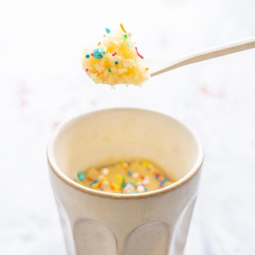 A spoon of vanilla cake decorated with sprinkles being held above a coffee mug