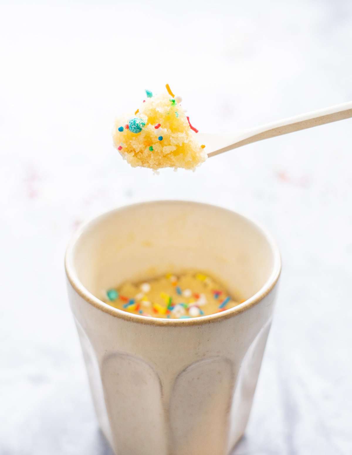 A spoon of vanilla cake decorated with sprinkles being held above a coffee mug