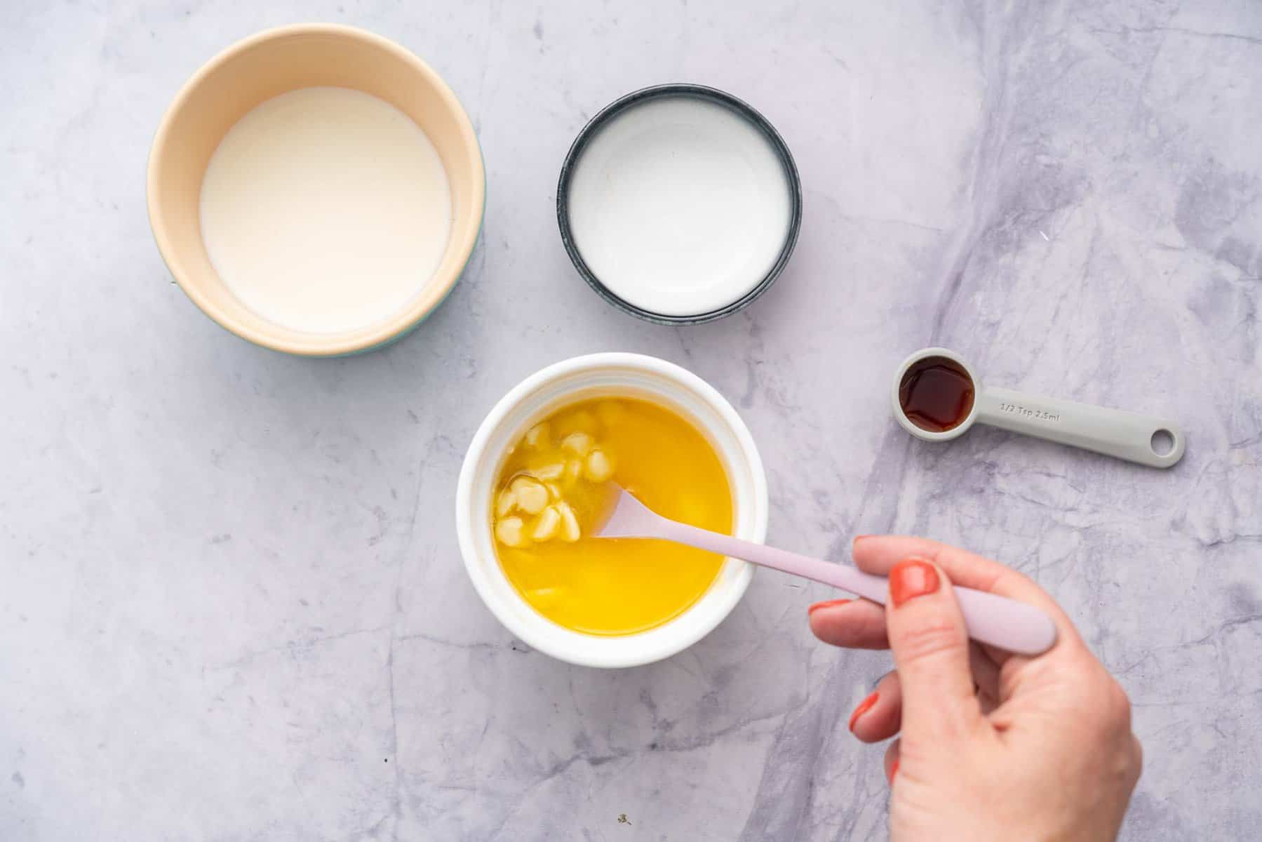 Ramekin showing the white chocolate and melted butter in a mug being mixed with a spoon next to ½ teaspoon of vanilla essence and emptied ramekins beside 