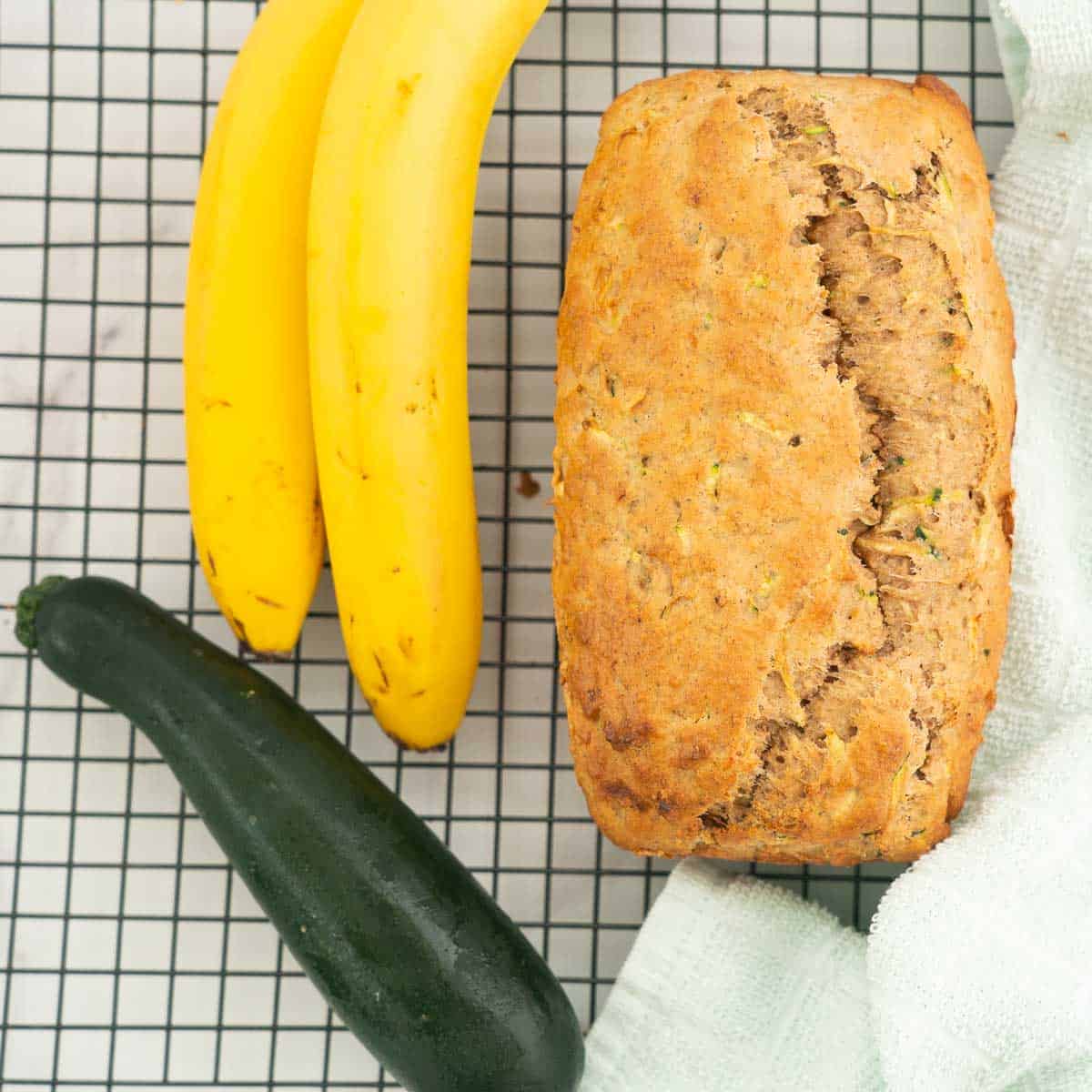 A loaf of banana bread resting on a cooling rack next to 2 bananas and a zucchini. 