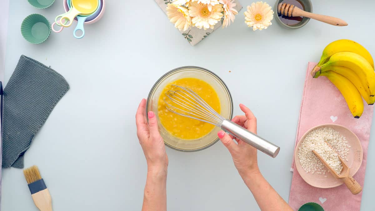 A glass mixing bowl filled with eggs and mashed bananas with a whisk
