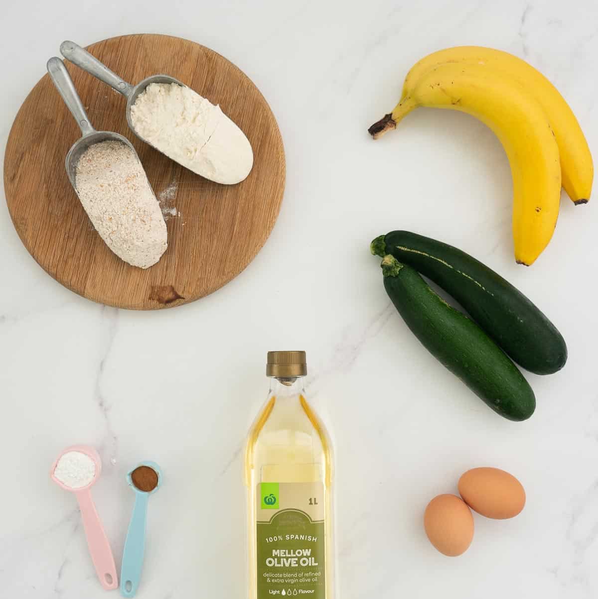 The ingredients to make banana zucchini bread laid out on a white marble bench top. 