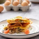An omelet filled with bacon, mushrooms tomatoes on a marble bench top in front of a carton of eggs