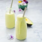 Two glass jars filled with Avocado Smoothie sitting on a bench with metal straws in each one as well as half an avocado and a small bowl of edible flowers in behind them.