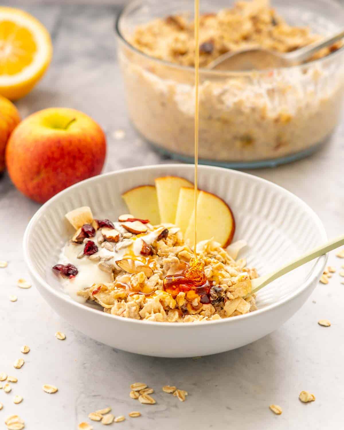 A dessert bowl with a serving of Bircher Muesli and sliced apples and drizzled honey sitting on the bench with a large serving dish in the background filled with Bircher Muesli in the background next to some red apples.