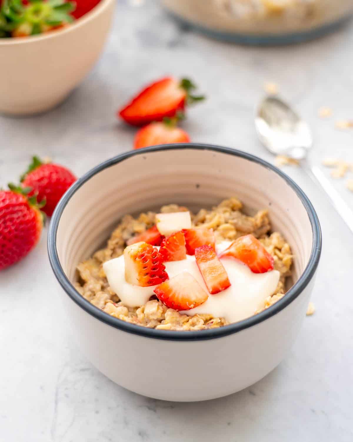 A serving of Bircher Muesli in a small ceramic dessert bowl with a tablespoon of greek yoghurt sitting on the bench amongst scattered strawberries and oats