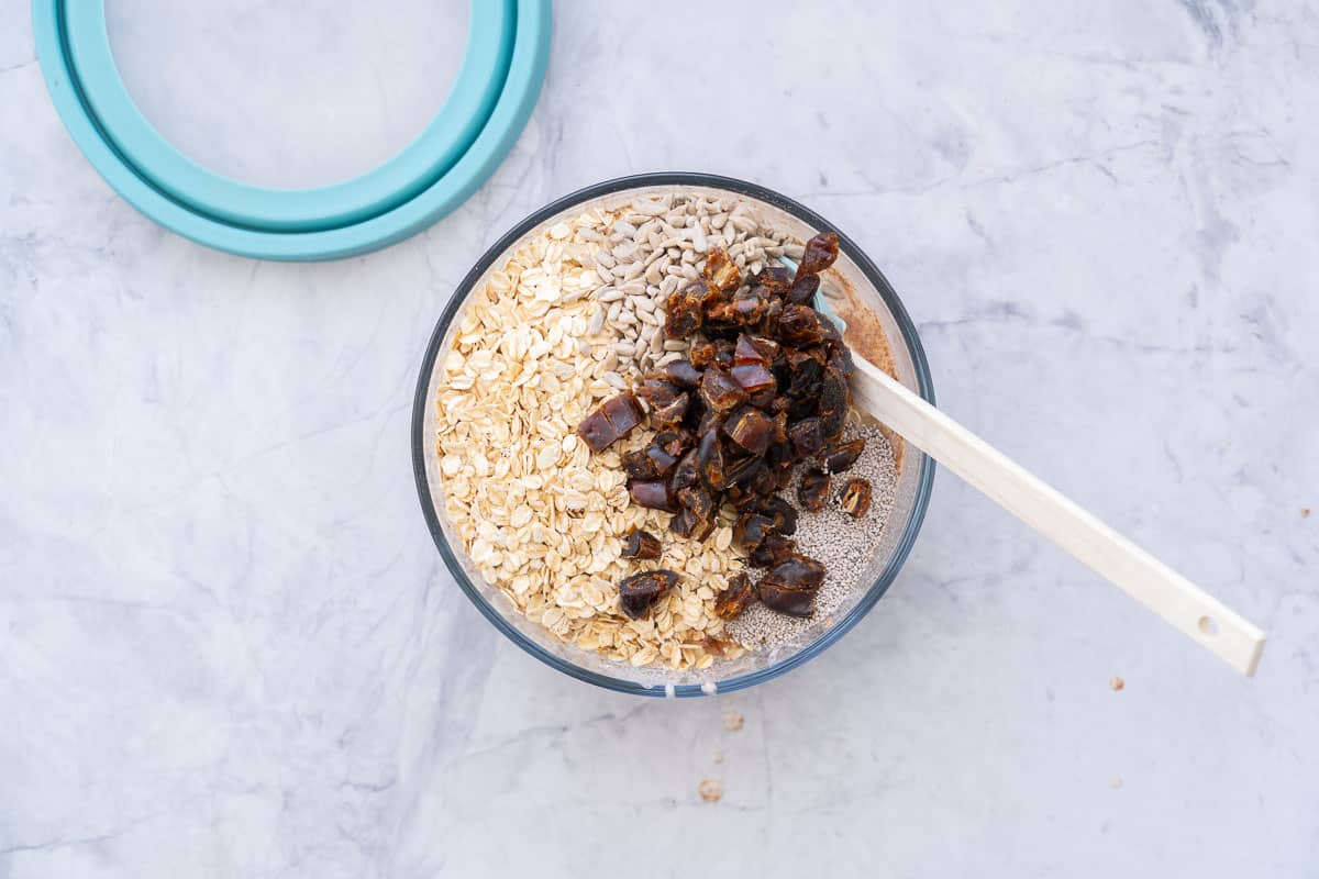 A wooden spoon stirring in the rolled oats, sunflower seeds, grated apples, dates, sunflower seeds and chia seeds into the wet ingredients in a bowl on the bench