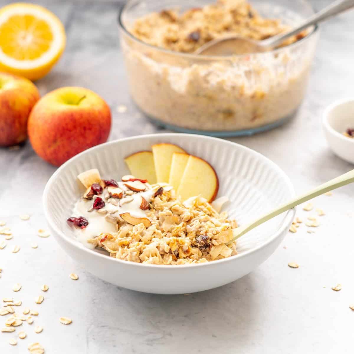 A dessert bowl with a serving of Bircher Muesli and sliced apples and drizzled honey sitting on the bench with a large serving dish in the background filled with Bircher Muesli in the background next to some red apples.
