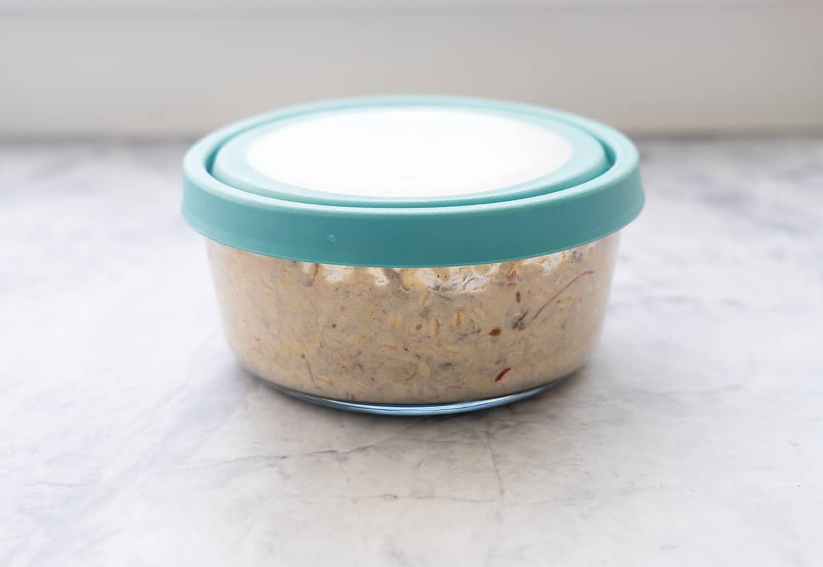 A large glass bowl filled with Bircher Muesli with the lid placed on sitting on the bench