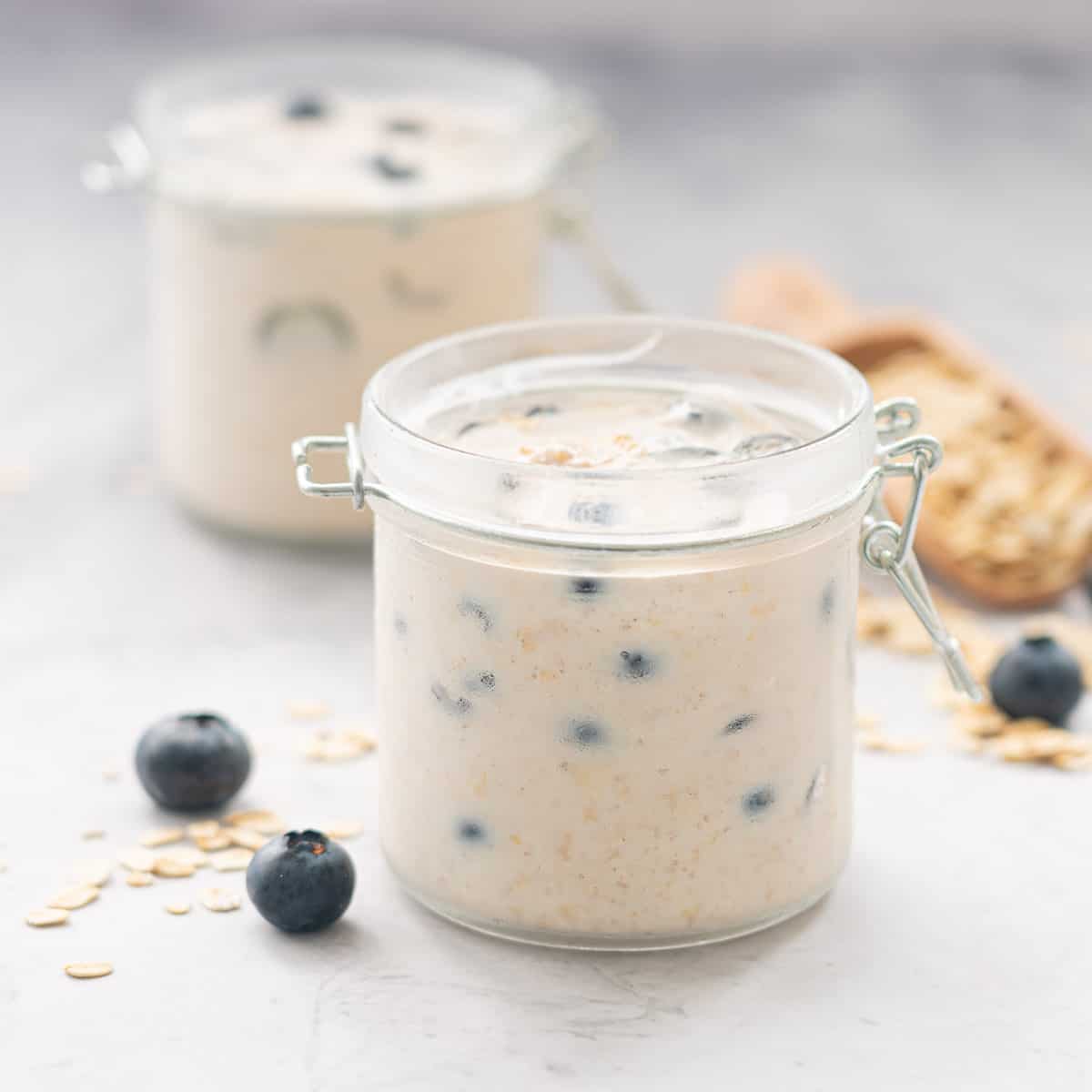 Two jars filled with Blueberry overnight oats sitting on a bench next to scattered blueberries and rolled oats 