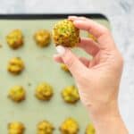 Cooked golden and crisp broccoli bites on a lined baking tray sitting on a bench with a hand holding one above them.